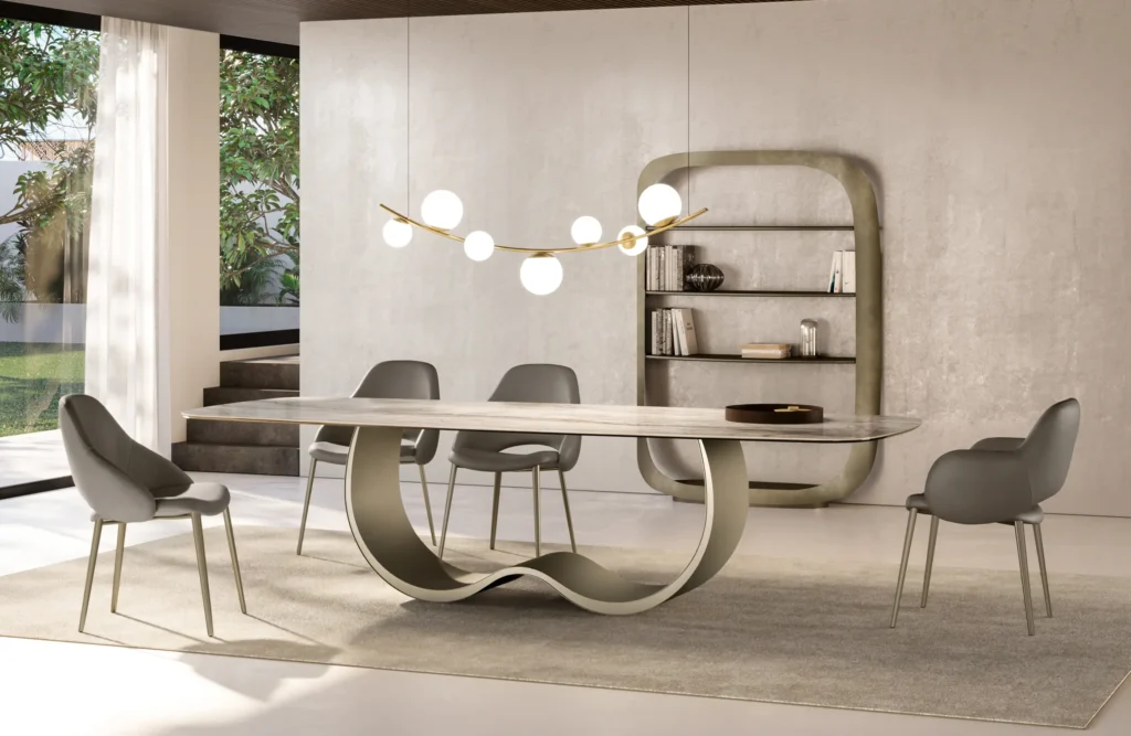 Une salle à manger moderne comprend une table élégante avec une base ondulée et quatre chaises grises. Un lustre unique avec des lumières sphériques est suspendu au-dessus. La pièce dispose d'une grande fenêtre, de murs clairs, d'une étagère minimaliste et d'une vue extérieure. Meubles Barret Bordeaux