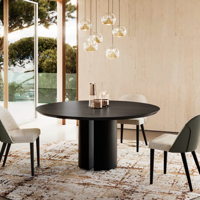 Une salle à manger contemporaine met en valeur la table ronde noire ALICE accompagnée de quatre chaises, posées sur un tapis à motifs, avec des suspensions au-dessus. De grandes fenêtres encadrent des vues panoramiques sur les arbres et l'extérieur, renforçant une ambiance lumineuse et naturelle. Meubles Barret Bordeaux