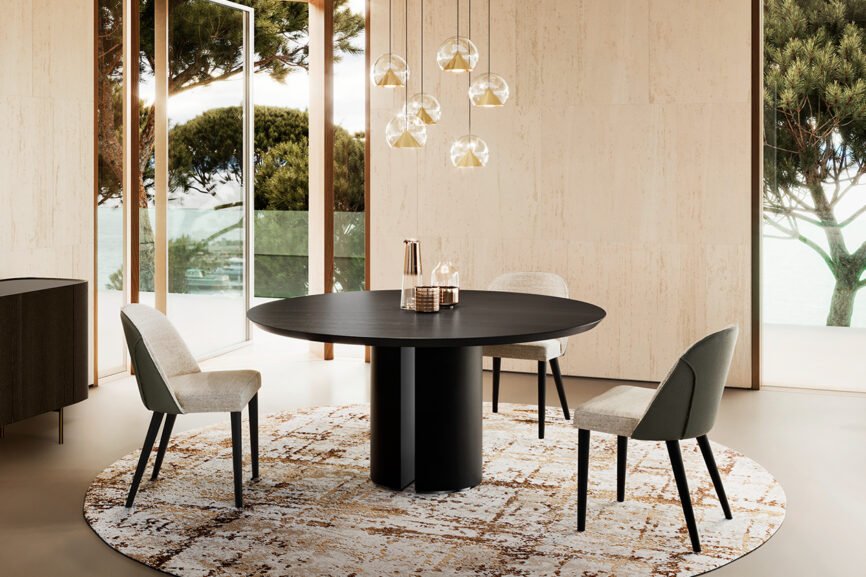 Une salle à manger contemporaine met en valeur la table ronde noire ALICE accompagnée de quatre chaises, posées sur un tapis à motifs, avec des suspensions au-dessus. De grandes fenêtres encadrent des vues panoramiques sur les arbres et l'extérieur, renforçant une ambiance lumineuse et naturelle. Meubles Barret Bordeaux