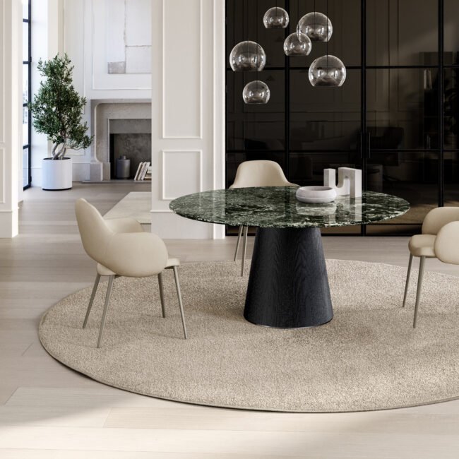 Une salle à manger élégante est dotée d'une table ronde en marbre accompagnée de quatre chaises beiges et d'un tapis rond. Des luminaires contemporains sont suspendus au-dessus, renforçant l'ambiance. Le miroir EMILY orne le mur, ajoutant de l'élégance à l'espace, tandis qu'une grande plante près de la fenêtre et une cheminée en arrière-plan complètent le cadre sophistiqué. Meubles Barret Bordeaux