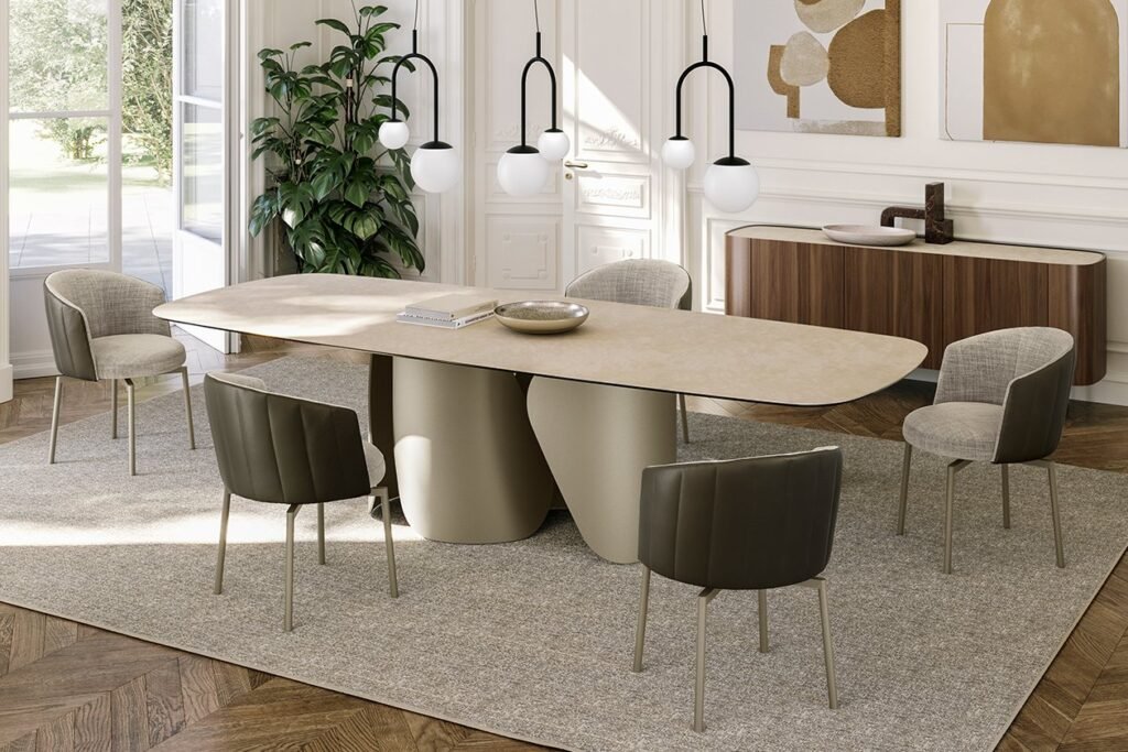 Une salle à manger contemporaine met en valeur la table JANE pieds métal avec son design ovale élégant et sa base distinctive, accompagnée de six chaises rembourrées moelleuses. Un buffet élégant et des œuvres d'art abstraites ornent le mur, tandis que trois luminaires suspendus flottent gracieusement au-dessus de la table. Pour compléter l'ambiance, une grande plante se dresse à proximité, avec une fenêtre offrant une vue sur la verdure luxuriante à l'extérieur. Meubles Barret Bordeaux