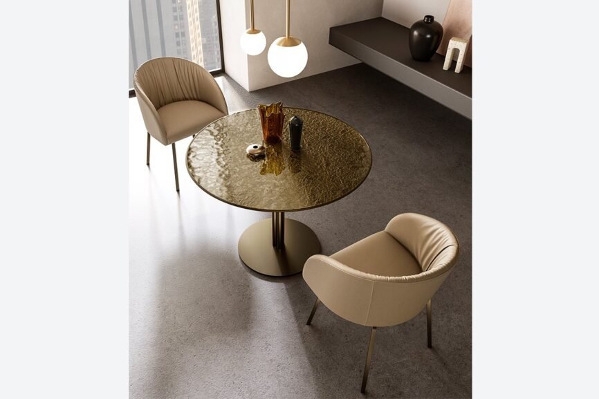 Une salle à manger contemporaine met en valeur la table VULCANO avec son plateau rond en verre texturé soutenu par un seul pied. Aux côtés de la table se trouvent deux chaises rembourrées beiges. Au-dessus, deux suspensions sphériques fournissent l'éclairage, tandis qu'une élégante étagère murale présente des objets décoratifs. Meubles Barret Bordeaux